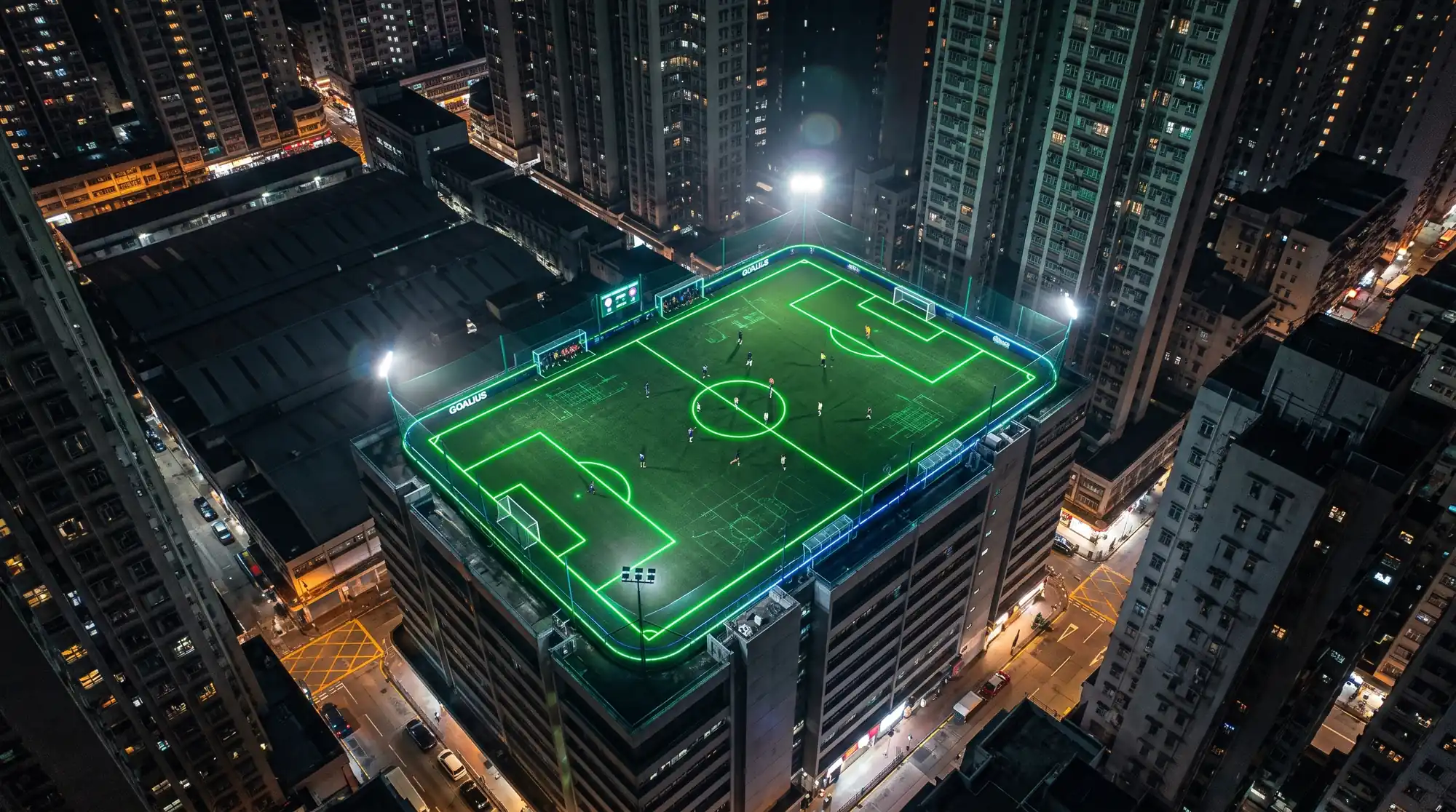 Vista aérea de una pista de fútbol 7 iluminada en la azotea de un edificio, rodeada de rascacielos de noche